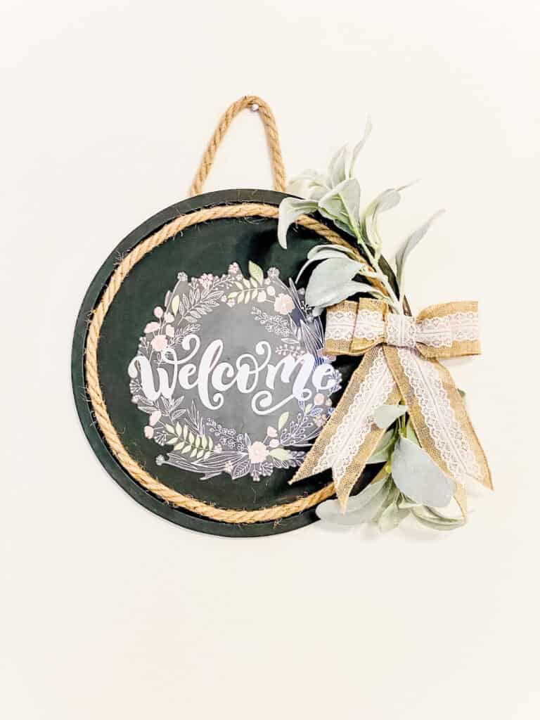 diy dollar tree craft with a painted pizza pan and welcome text attached to make a welcome sign.  A decorative plant and bow is on the side.