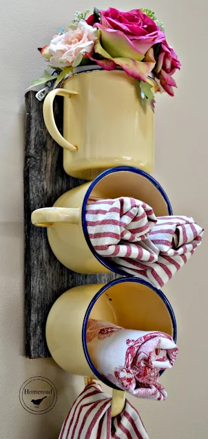 Three yellow enamel mugs attached to a wood board and hanging on a wall. The top cups is holding flowers and the bottom two are used as organizers for tea towels.